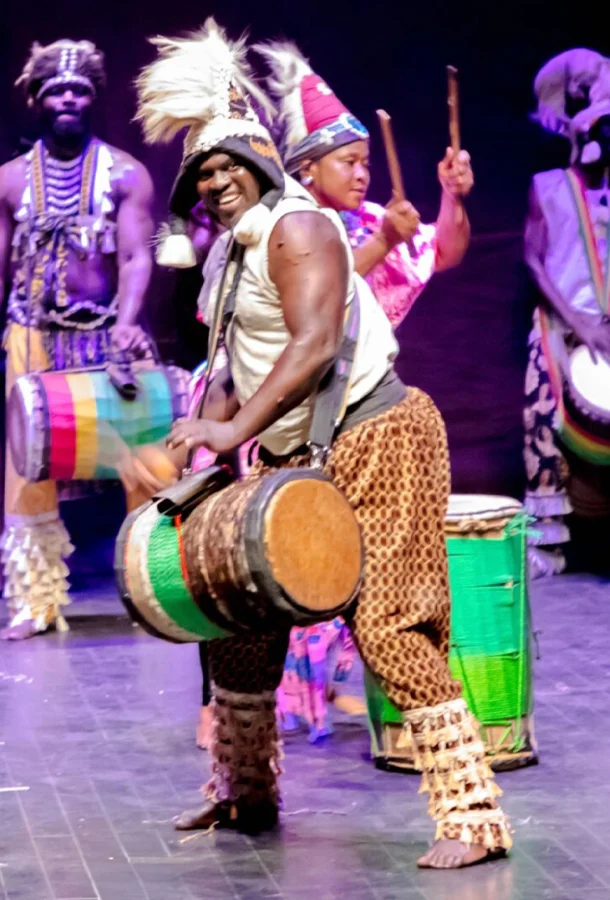 Man in colorful outfit drumming on stage with others.