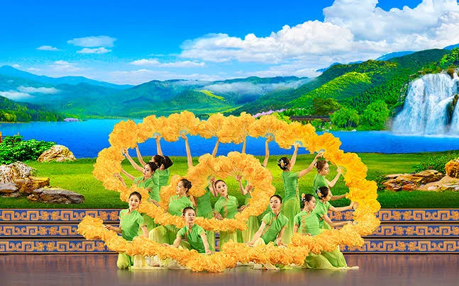 Dancers in bright green in a formation holding orange feathered fans in a swirl shape with a backdrop of Chinese lake and mountains.
