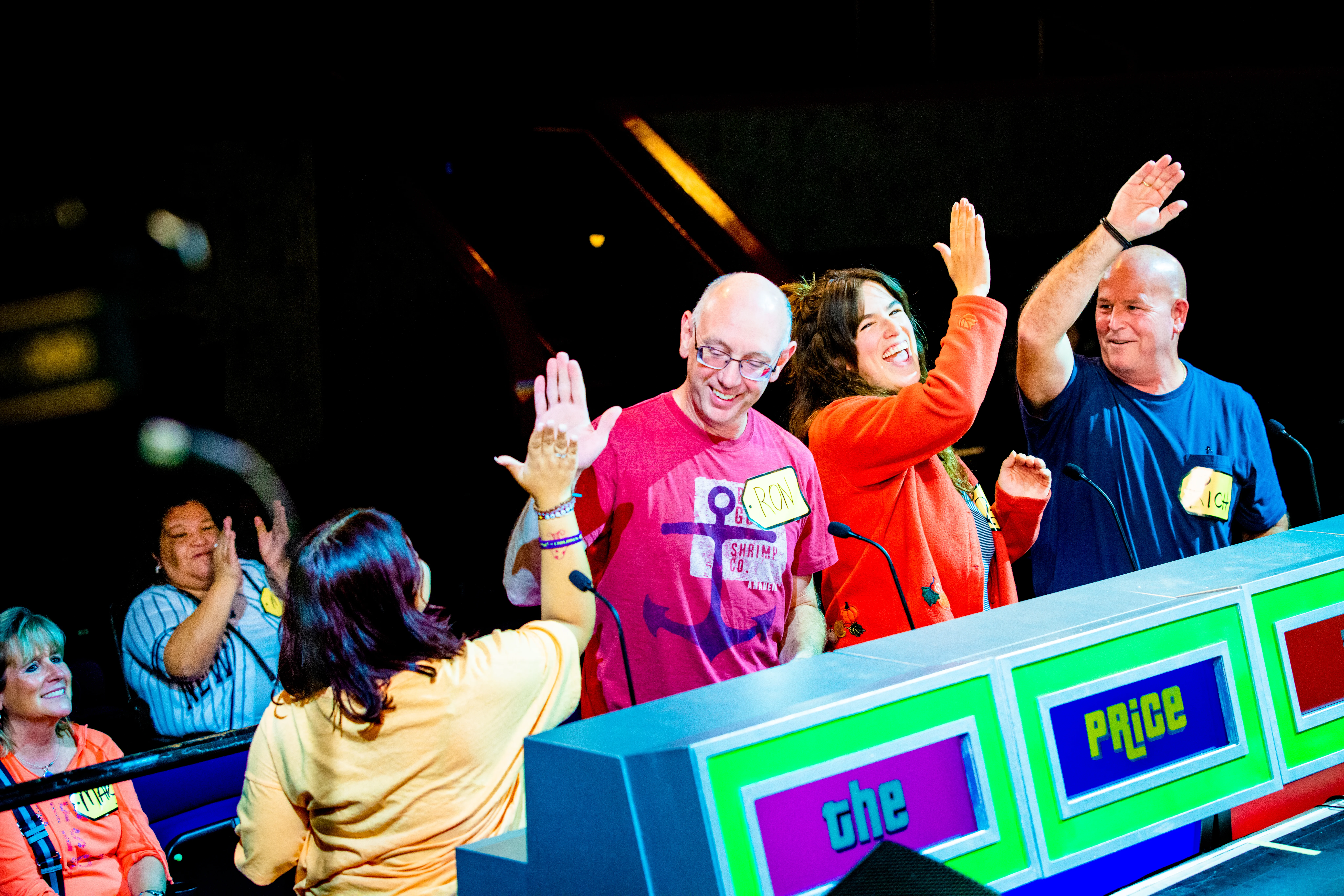 Group of people standing behind a colorful podium with the text 'the Price,' wearing name tags and raising their hands, participating in a game show 'The Price is Right'.