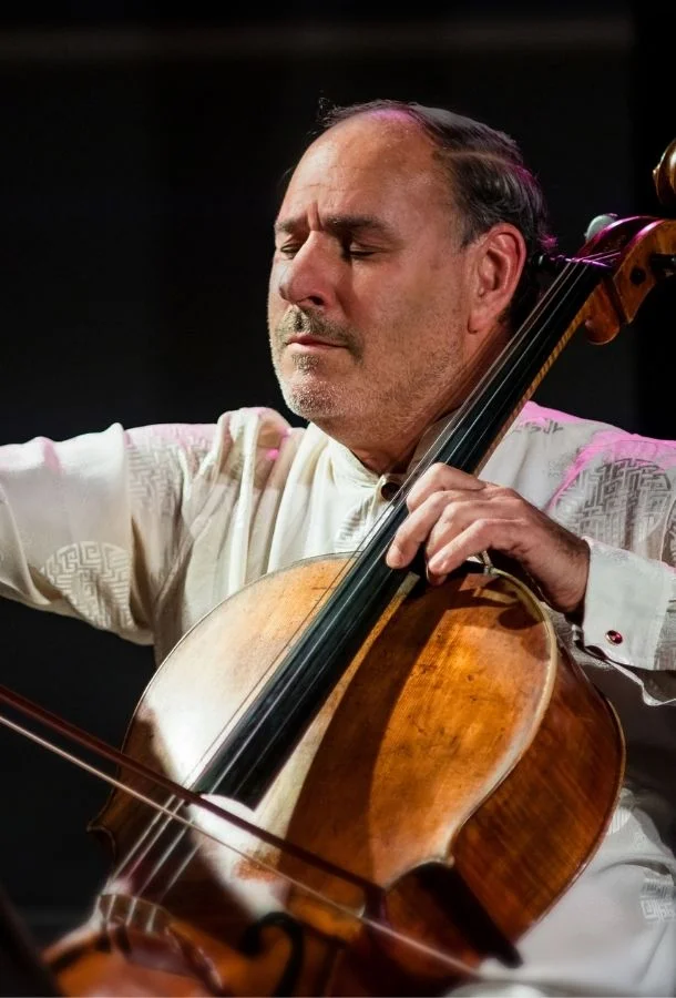 A person playing a wooden cello, wearing a light-colored shirt with patterned sleeves—one hand holding the bow, the other pressing the strings.