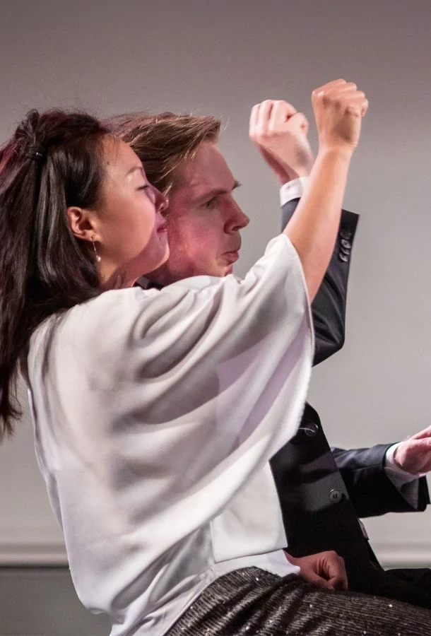 Two individuals with raising their arms—one wearing a white top with wide sleeves, the other in a dark suit—against a plain, neutral background.