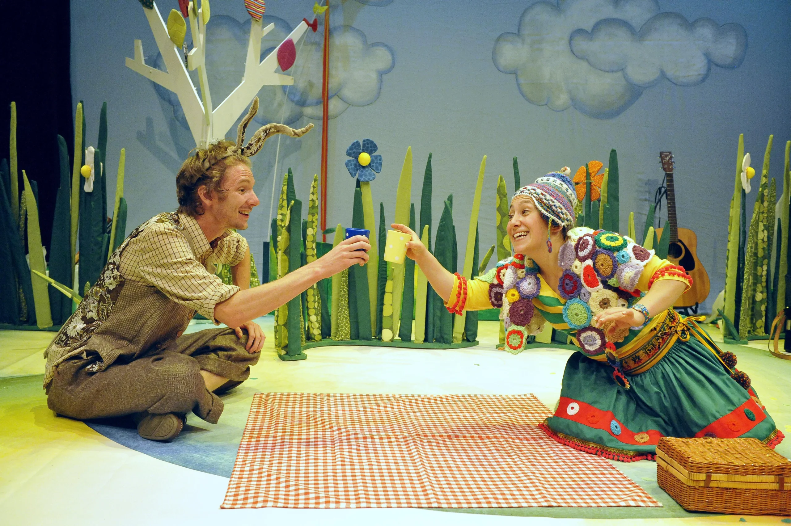 Two costumed people having a picnic on a checkered blanket in a whimsical stage setting with plants, props, and decorations.