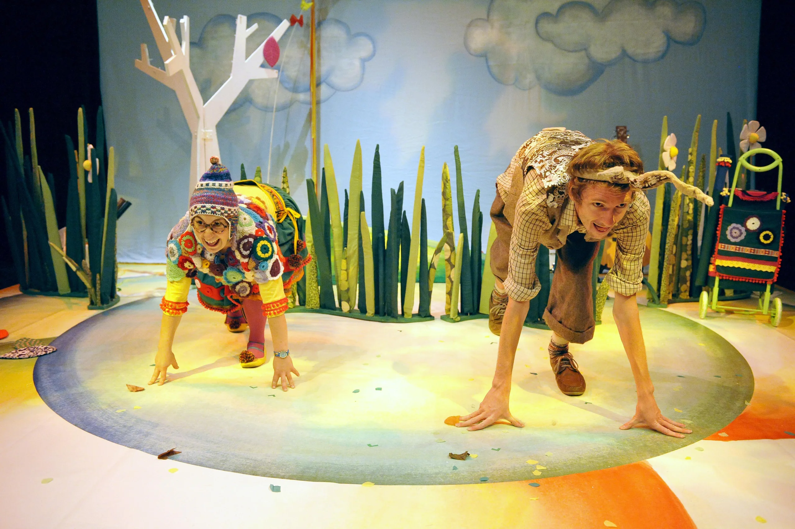 Two individuals crawling on a colorful stage with tall green plants, a white tree with red leaves in the background, and a colorful chair on the right side.
