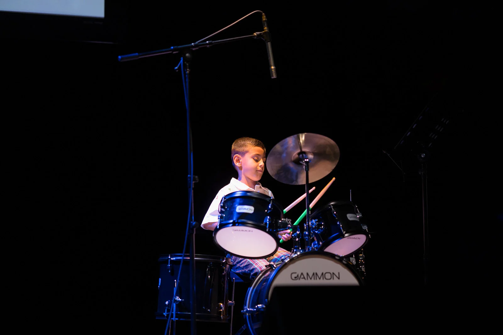 Young student playing drum set on dark lit stage.	