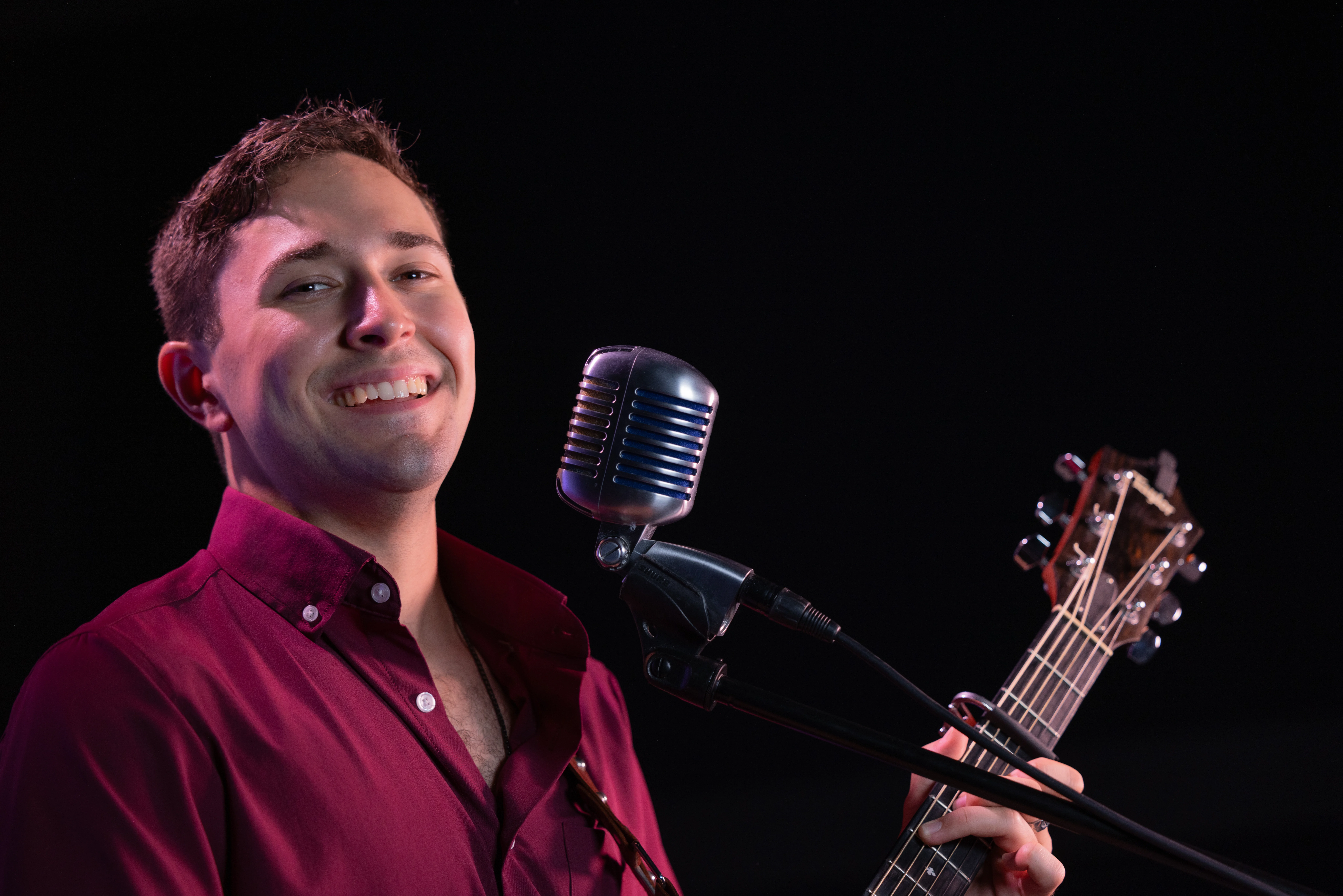Mike Daly sings into microphone with guitar smiling.