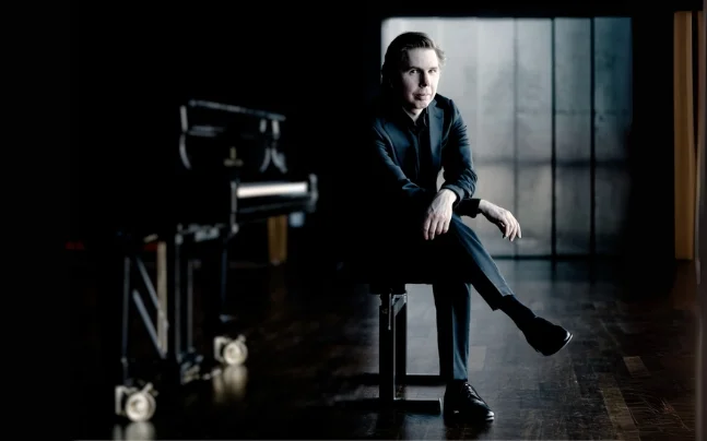 Double Forte Chamber Music Society of Palm Beach Juho Pohjonen in a suit sitting on a piano bench in low lighting.