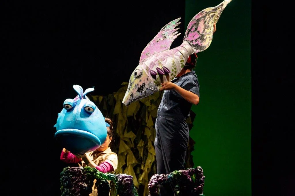 Two people operating large, colorful fish puppets on a stage—one blue with a round face and large eyes, the other pink and white with wing-like fins—set against a dark background with green lighting and underwater plant props in the foreground.
