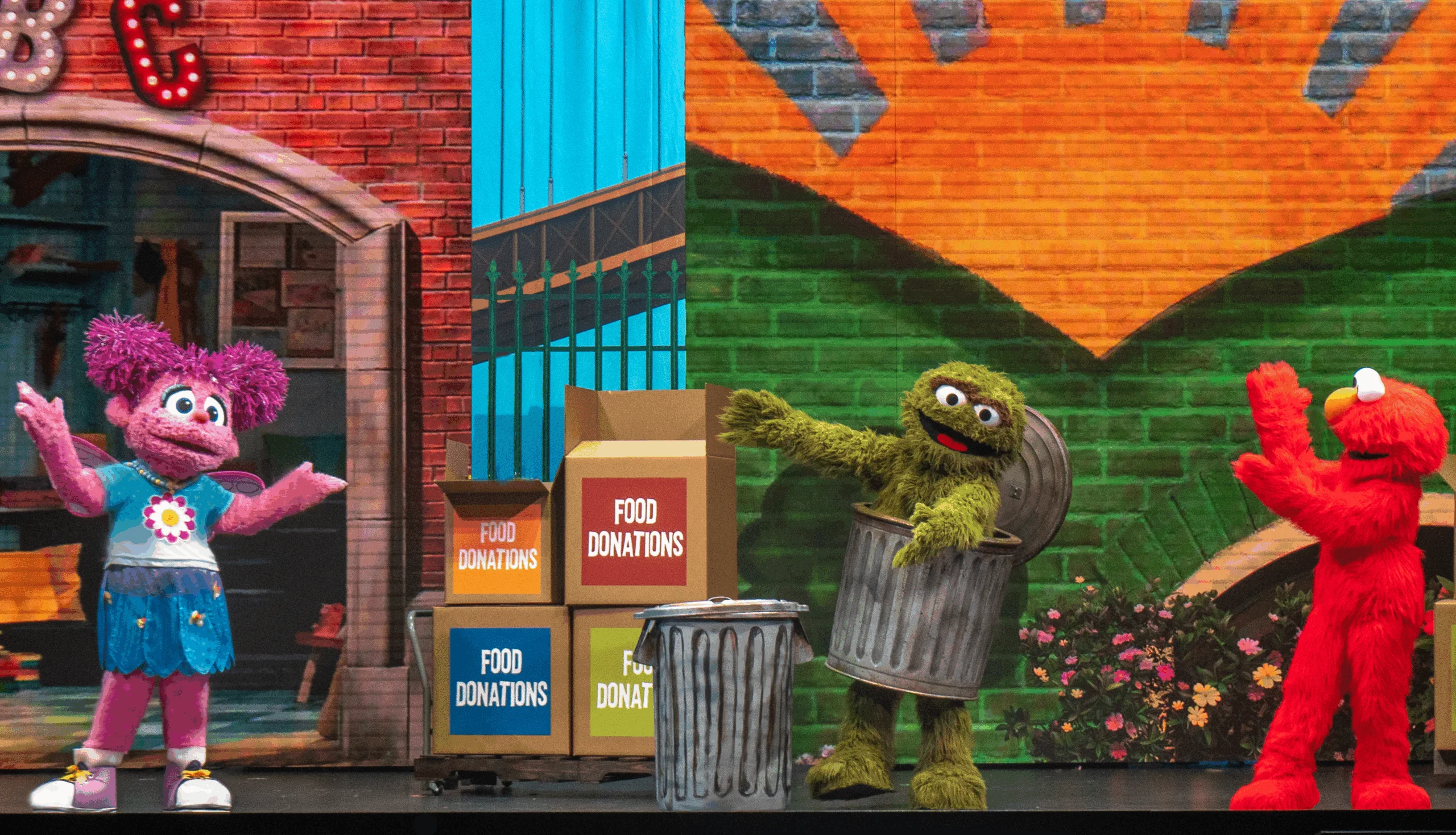 Abby Caddaby, Oscar the Grouch and Elmo on stage with food donation boxes on a cart.
