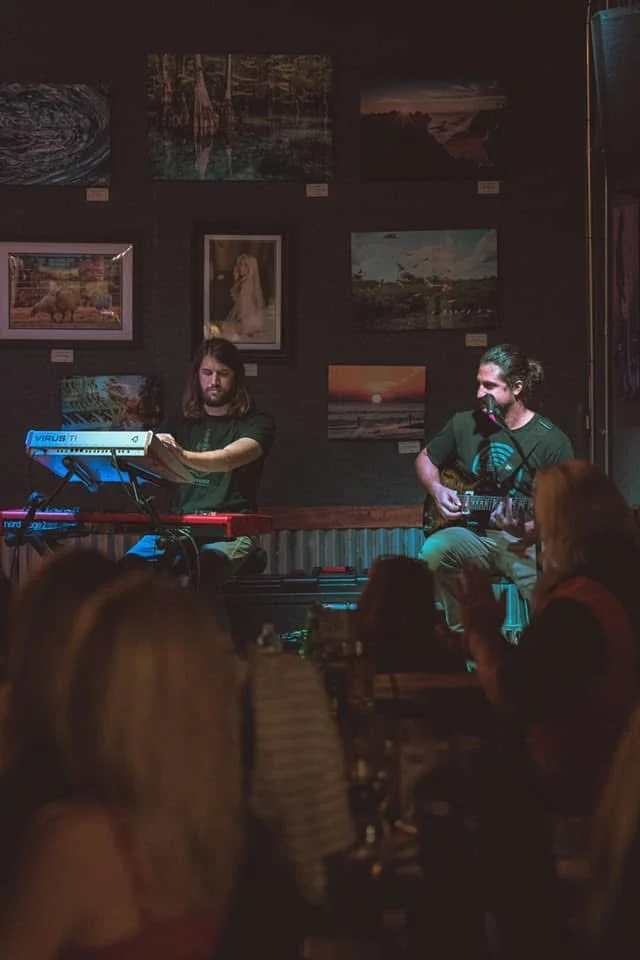 Two men performing in a dark lit corner in front of hanging artwork.