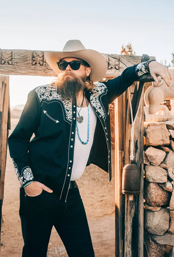 A person with a long beard and sunglasses stands outdoors, leaning against a wooden gate. They wear a wide-brimmed hat, a black jacket with white embroidered details, a white shirt, and dark pants. The background shows a desert landscape with rocks and sparse vegetation.