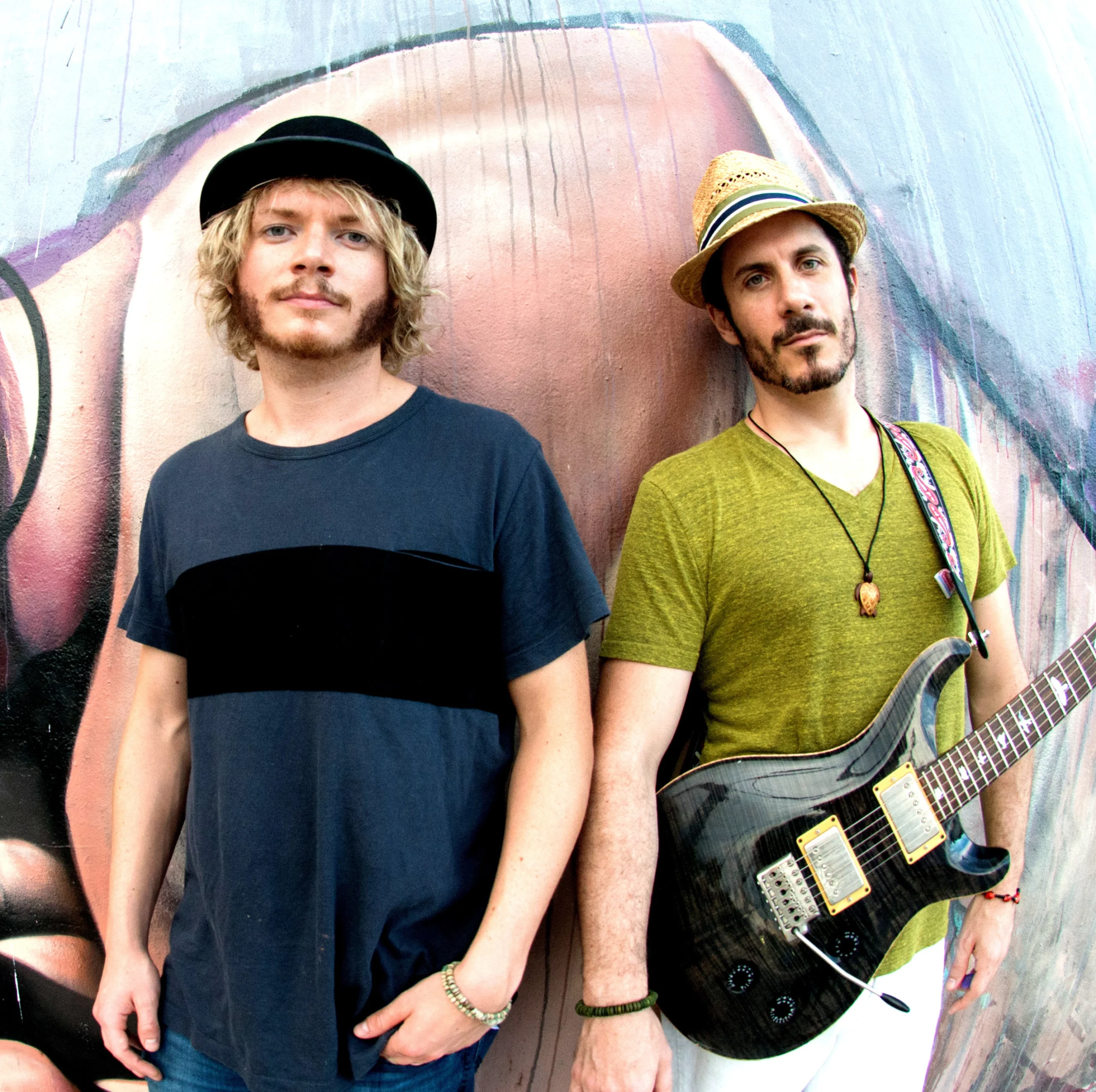 Spider Cherry band pose in front of mural with one in green wearing a guitar and other one in black shirt.