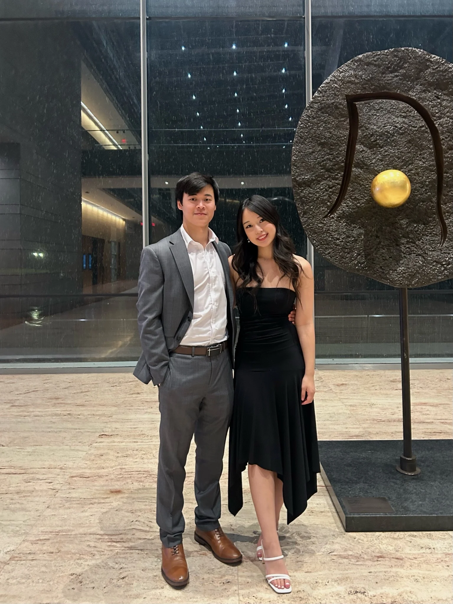 Two people in formal attire posing at an art gallery with a modern sculpture in the background.