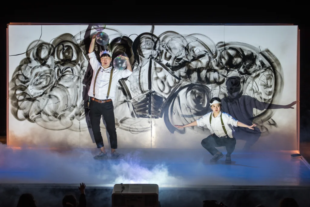 Two performers interacting with a large scale “doodle” in the background.