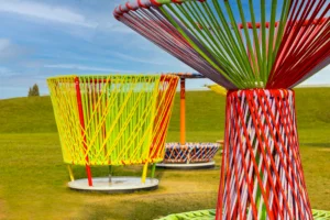 Large outdoor art installation featuring oversized spinning tops made of colorful woven cords. The tops are arranged on a grassy field under a blue sky. The closest top is red, green, and white, while others in the background display bright yellow, orange, and multicolor patterns.