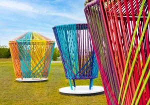 Large outdoor art installation featuring oversized spinning tops made of colorful woven cords. The tops are arranged on a grassy field under a blue sky. The closest top is red, green, and white, while others in the background display bright yellow, orange, and multicolor patterns.