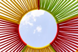 Outdoor art installation featuring oversized spinning tops made of colorful woven cords. The closest top is red, green, bright yellow and multicolor patterns.