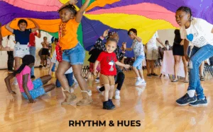 Rhythm & Hues. Children and adults playing under a large colorful parachute indoors on a polished wooden floor during a community activity.