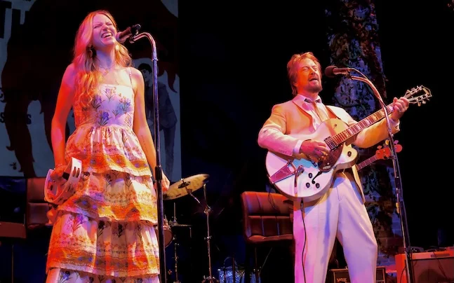 Two performers on stage singing and playing guitar.