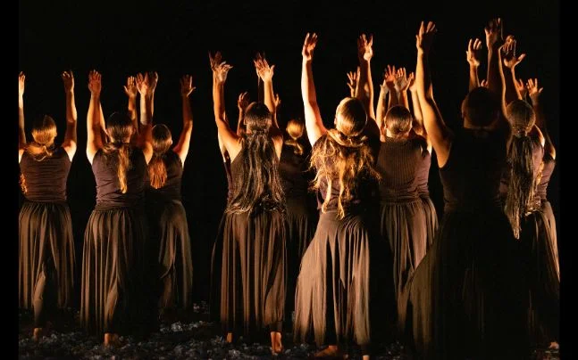 Group of dancers on stage wearing flowing black costumes, standing in a semi-circle with arms raised toward the light in a dramatic contemporary dance performance.