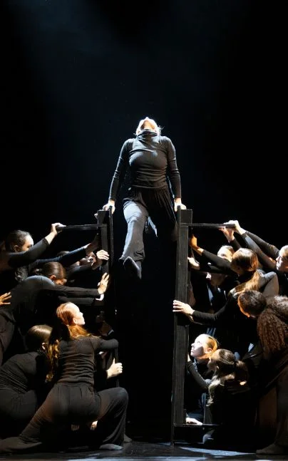 Dynamic stage performance featuring a group of dancers in black costumes surrounding a central performer elevated on a vertical structure under dramatic spotlight.