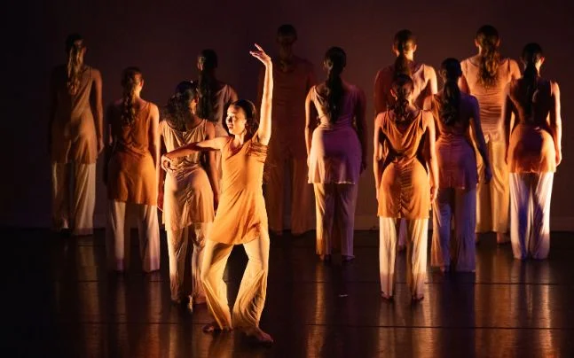 Contemporary dance performance featuring a group of dancers in coordinated peach and white costumes on a dimly lit stage, with one dancer striking an expressive pose in the foreground.