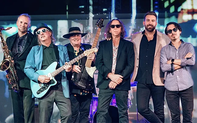 The 6 member of Expanding Man pose on stage. Some hold guitars or saxophone.