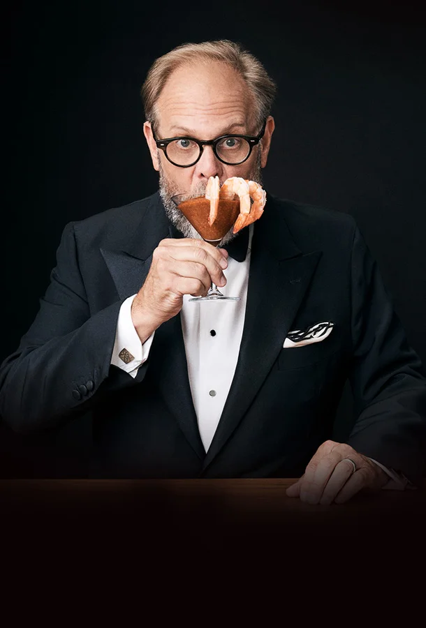 Alton Brown in formalwear raising a glass in a dramatic, studio-lit setting.