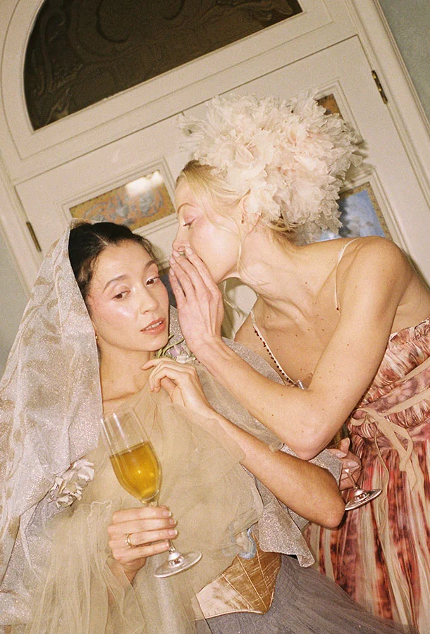 A woman in an elegant, feathered headpiece leans in to whisper to another festively dressed woman holding a champagne flute at a vintage‑style celebration.