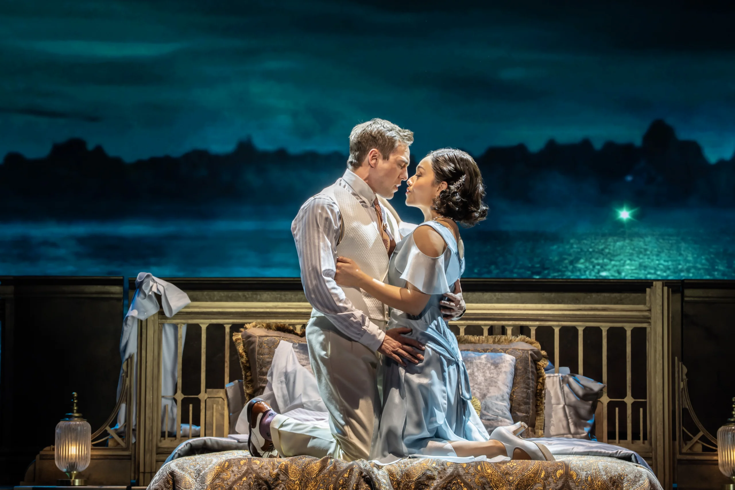 The Original West End cast singing while in a romantic embrace. A male presenting actor on the left and a female presenting actress on the right kneel on a luxurious bed while in elegant 1920s attire. As they gaze intently into each other’s eyes, the green light over the water shines behind them through the nighttime. Photo by Johan Persson.