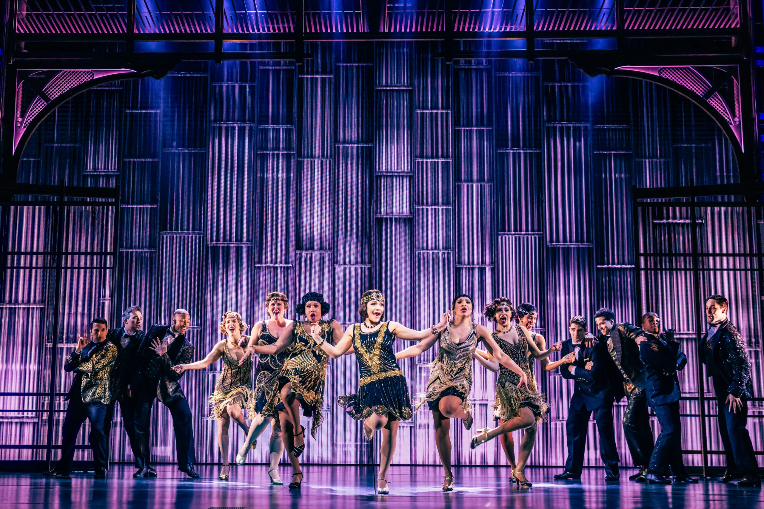 Noah J. Ricketts stands center stage in the Broadway production of The Great Gatsby, illuminated by a spotlight against a striking, blue-lit backdrop, with silhouetted cast members striking elegant poses in the background. Photo by Matthew Murphy and Evan Zimmerman.