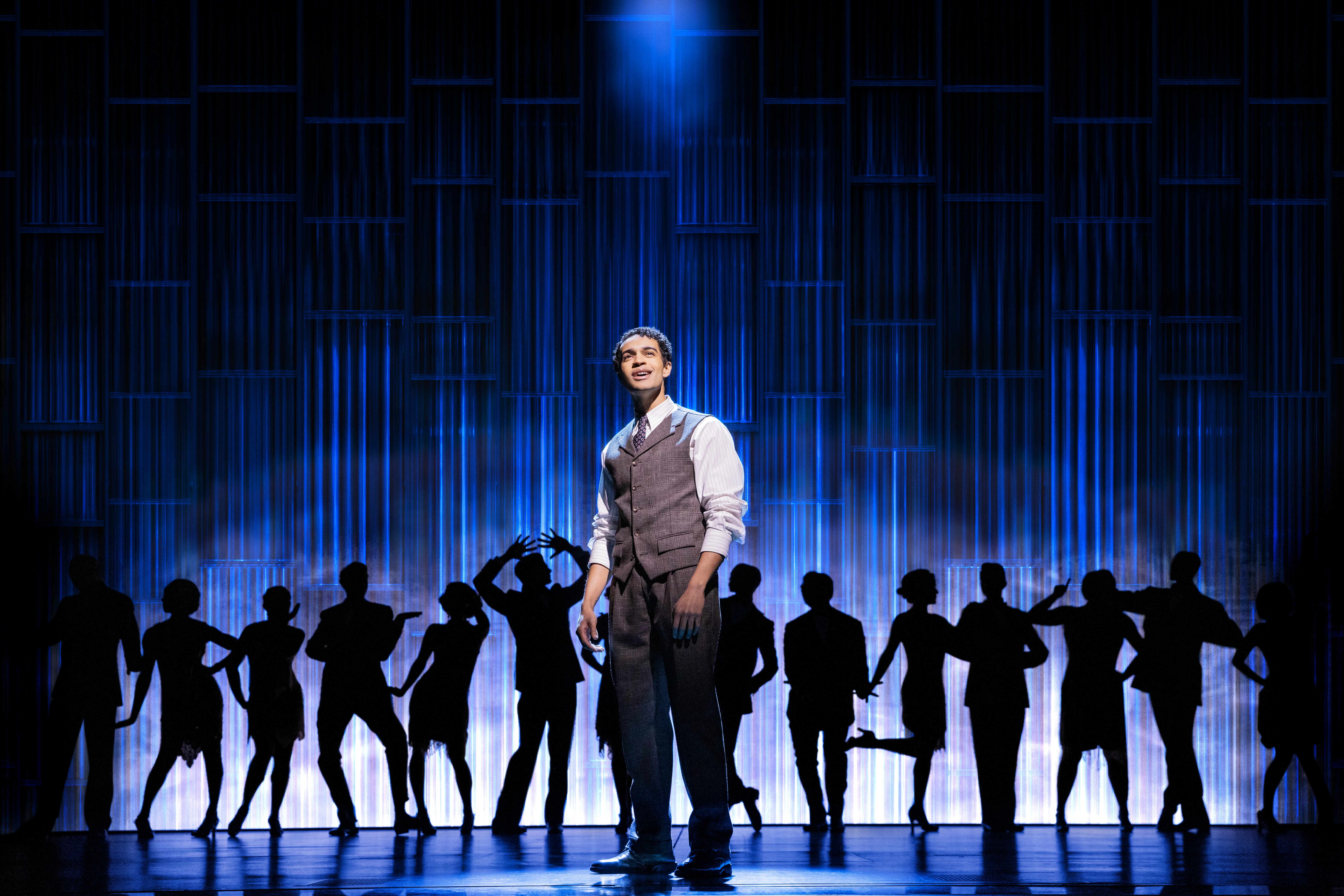 Noah J. Ricketts stands center stage in the Broadway production of The Great Gatsby, illuminated by a spotlight against a striking, blue-lit backdrop, with silhouetted cast members striking elegant poses in the background. Photo by Matthew Murphy and Evan Zimmerman.