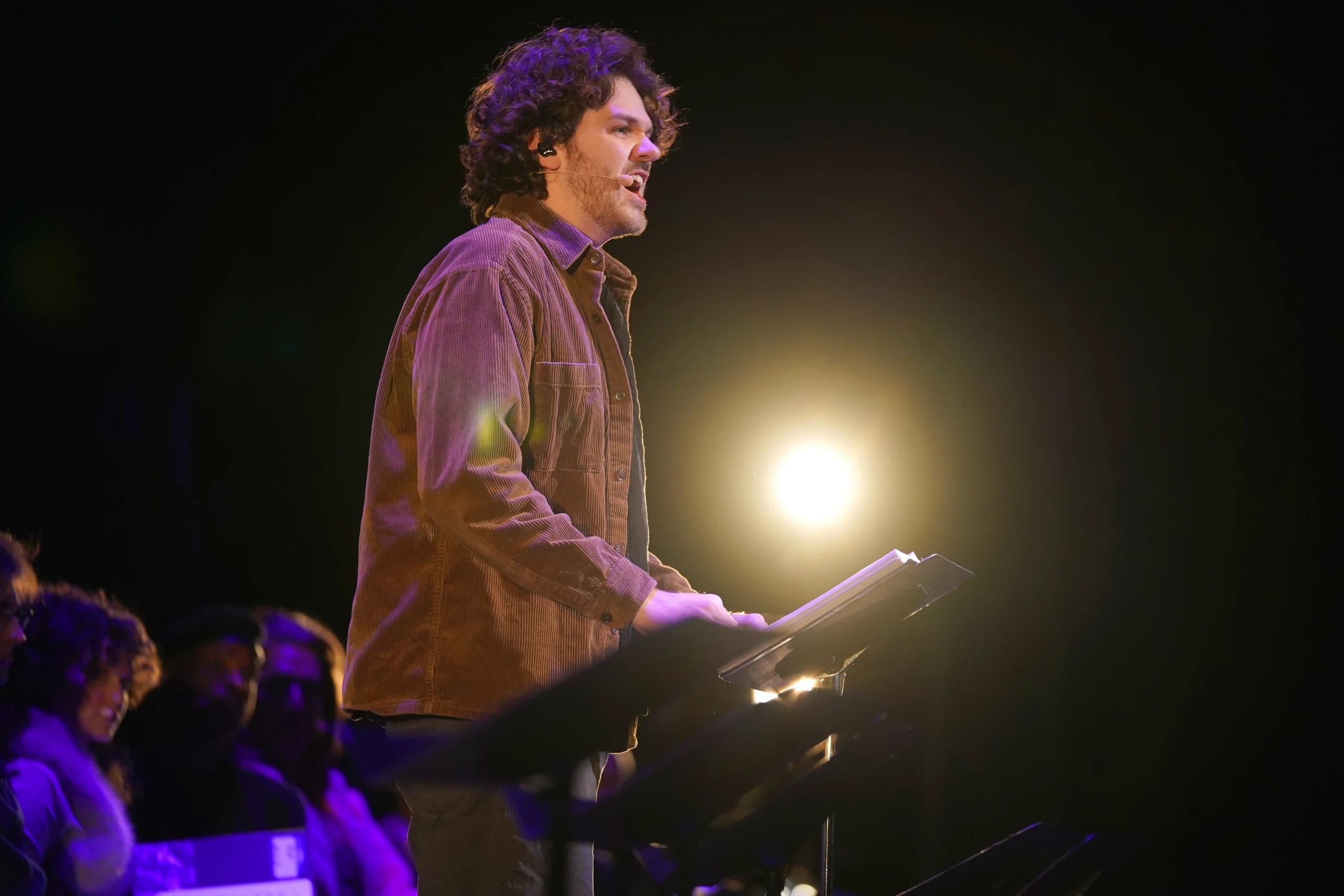 A performer stands at a music stand on a dimly lit stage, with a bright spotlight shining behind them and additional cast members seated off to the side.