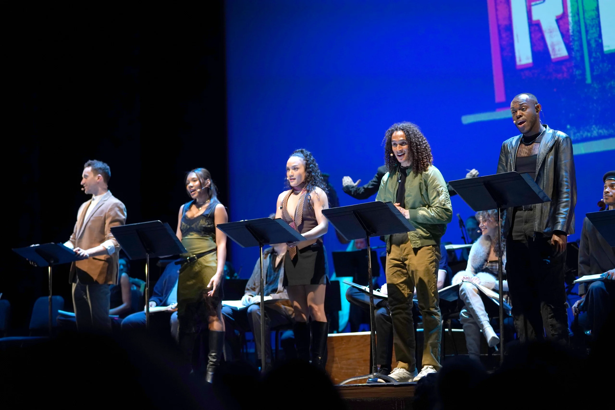 A group of performers stands onstage in front of music stands, illuminated by stage lighting, with musicians seated behind them. A large projected graphic is partially visible on the backdrop.
