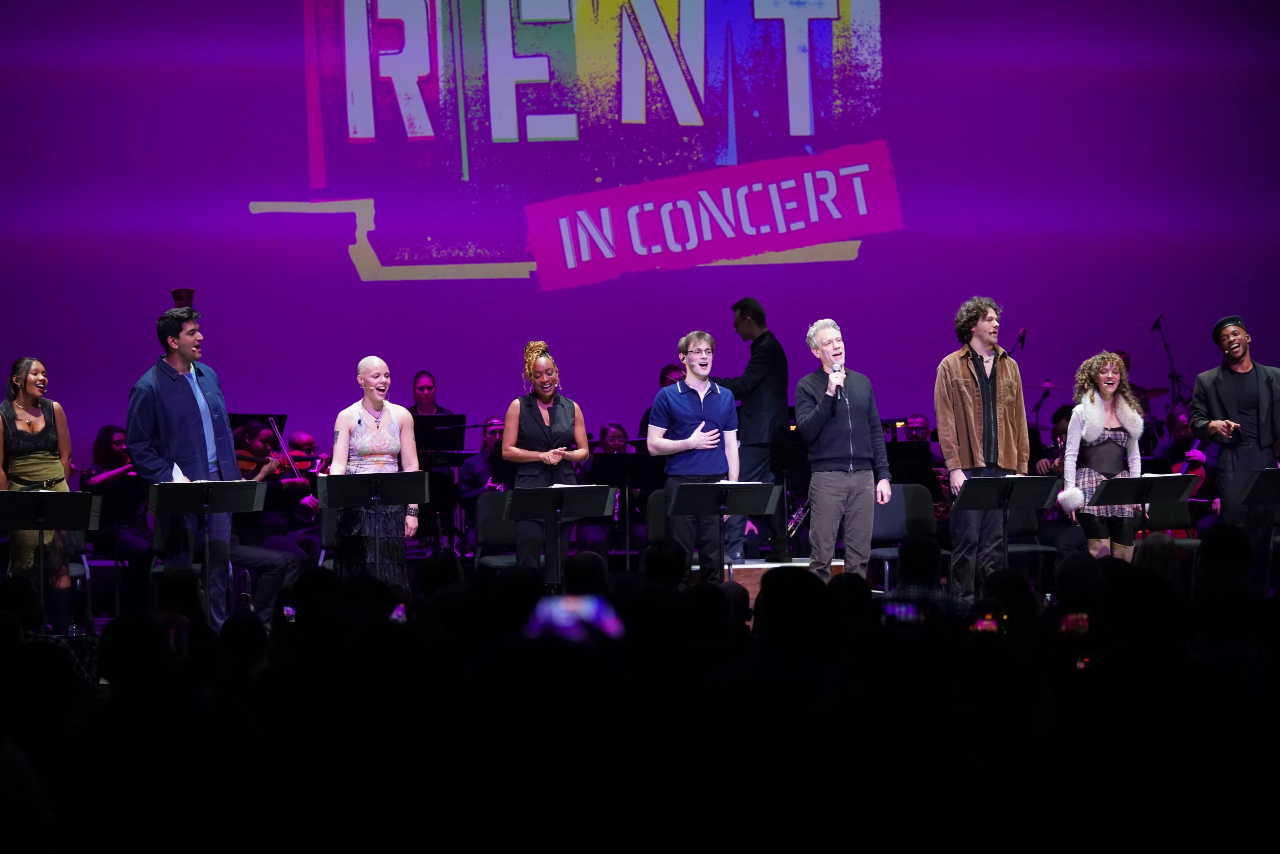 A full cast stands across the stage in front of music stands during a performance, with the backdrop displaying the text “RENT in Concert” in large colorful lettering.