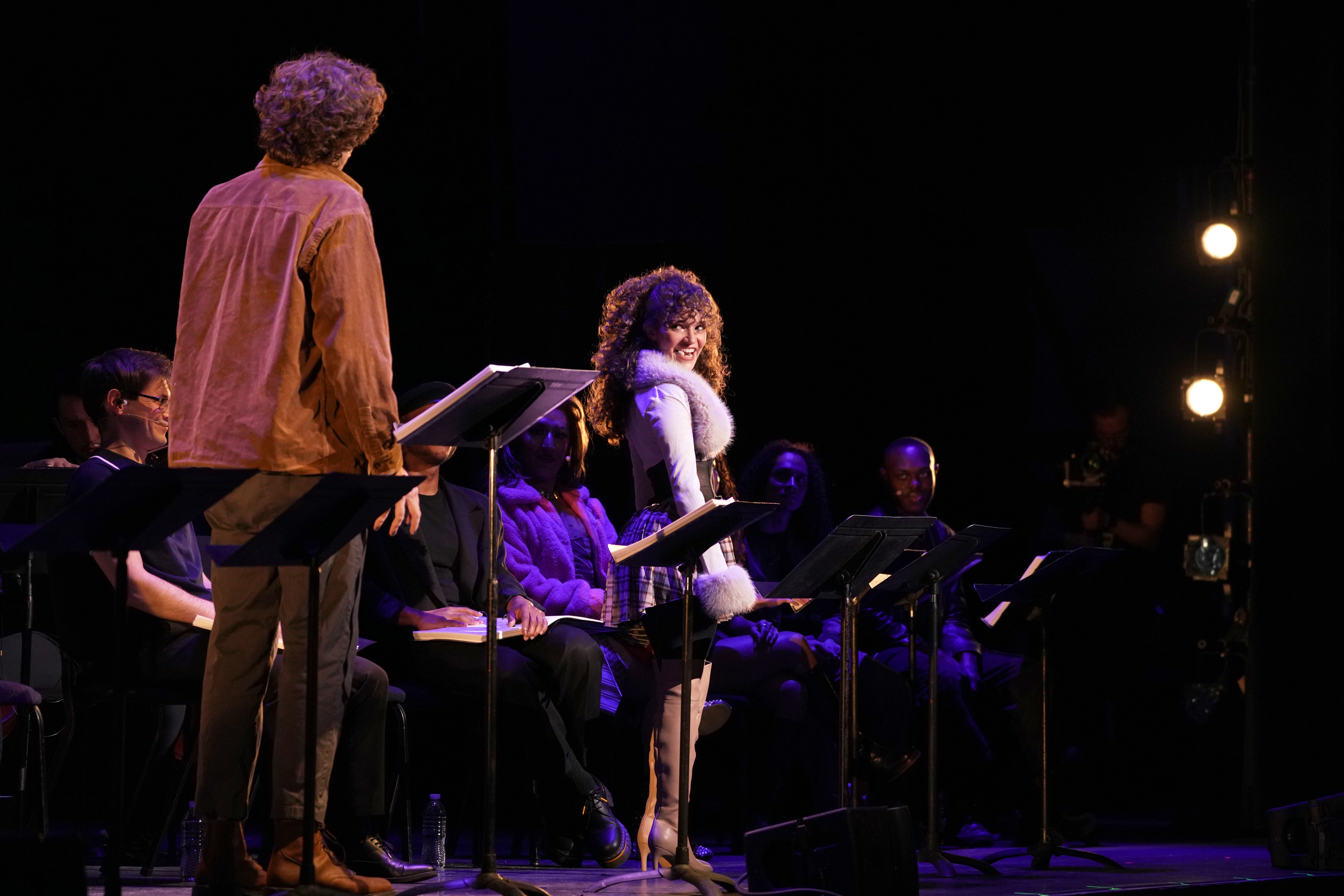 Two performers stand at music stands on a stage, facing each other during a staged reading, while additional cast members sit in chairs behind them under theatrical lighting.