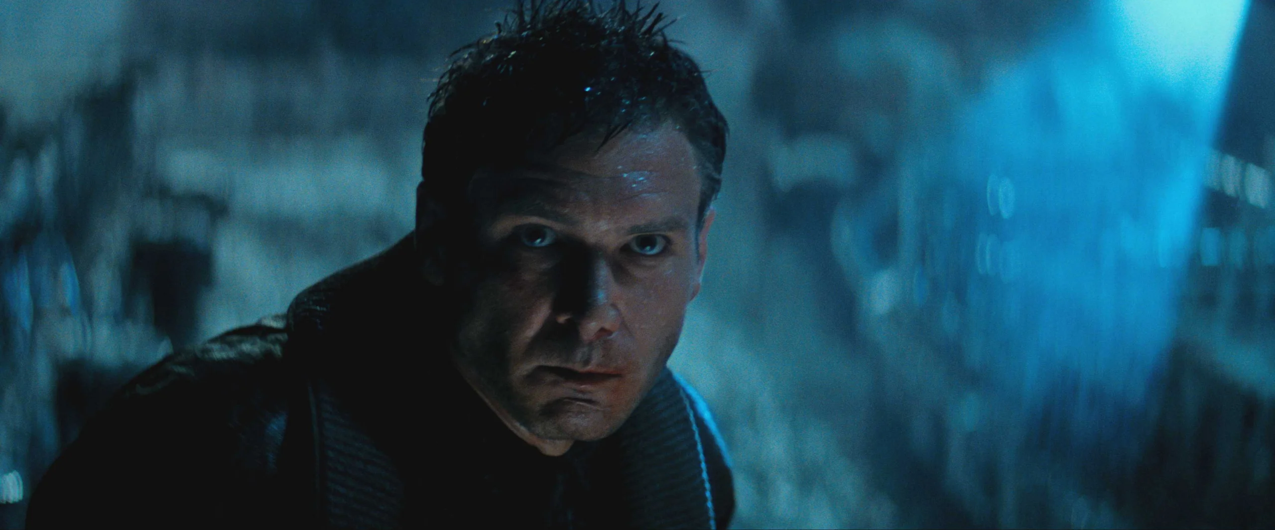 A dark, rainy, nighttime scene with a person standing in the foreground. The lighting is dim and cast in cool blue tones, with heavy rain falling around the figure. The background shows an industrial or urban environment with indistinct shapes, metallic structures, and soft blue illumination creating a moody, atmospheric setting.