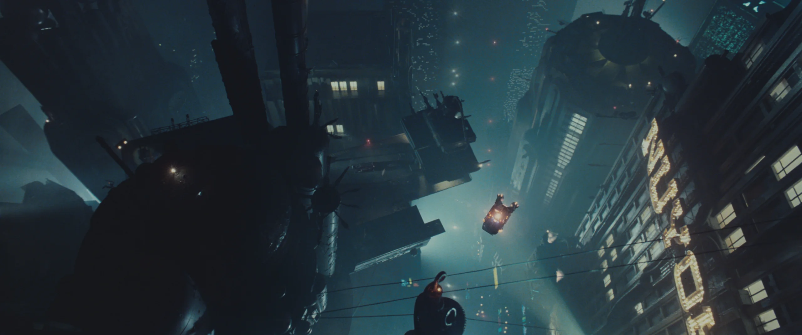 A futuristic cityscape at night, viewed from a dramatic tilted angle. Tall buildings with glowing windows and neon signage rise into the misty sky. Several flying vehicles move between the structures, leaving trails of light. The scene is filled with deep shadows, blue ambient light, and dense atmospheric haze, contributing to a cyberpunk aesthetic.
