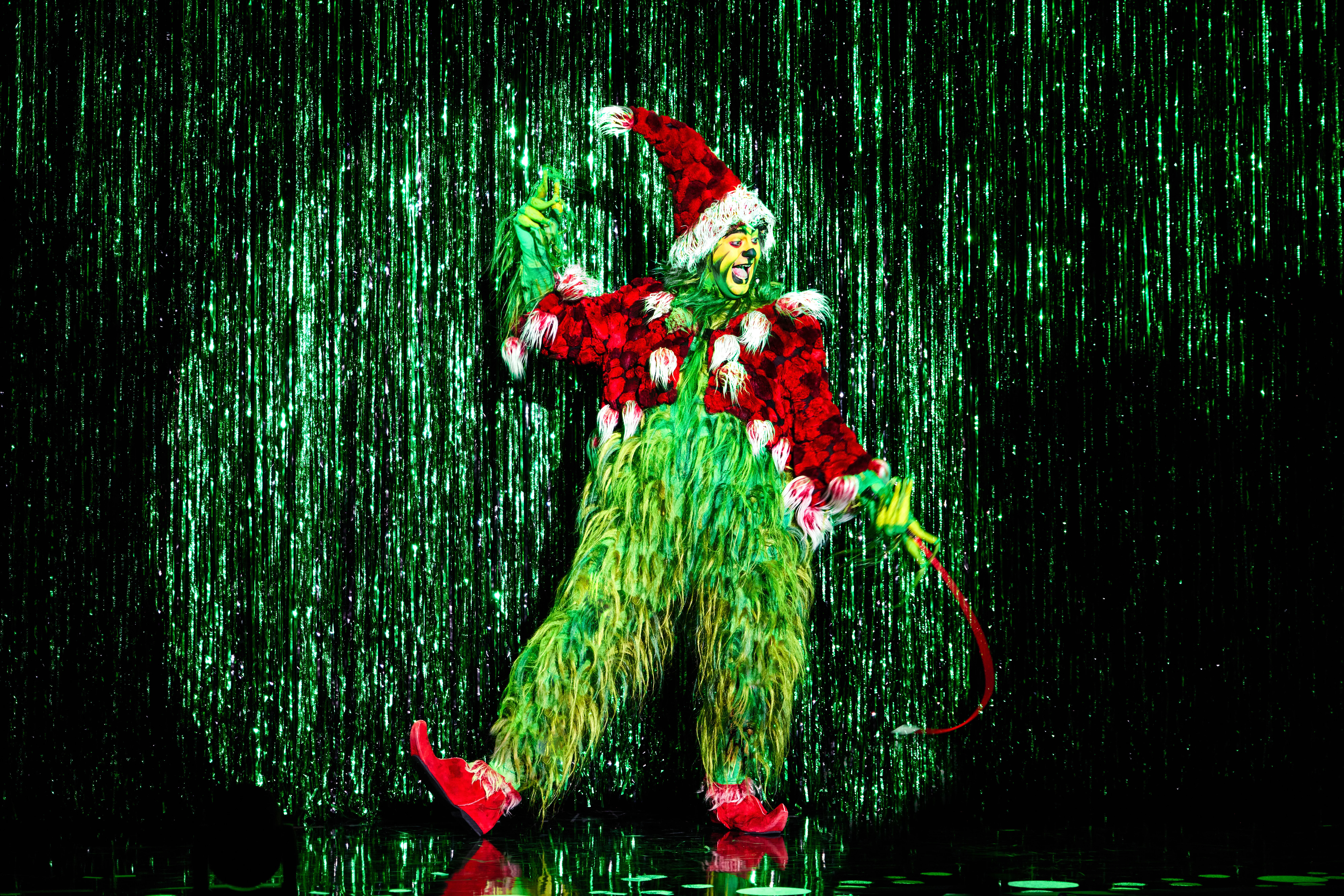 THE GRINCH in spotlight in front of shinny green curtain.