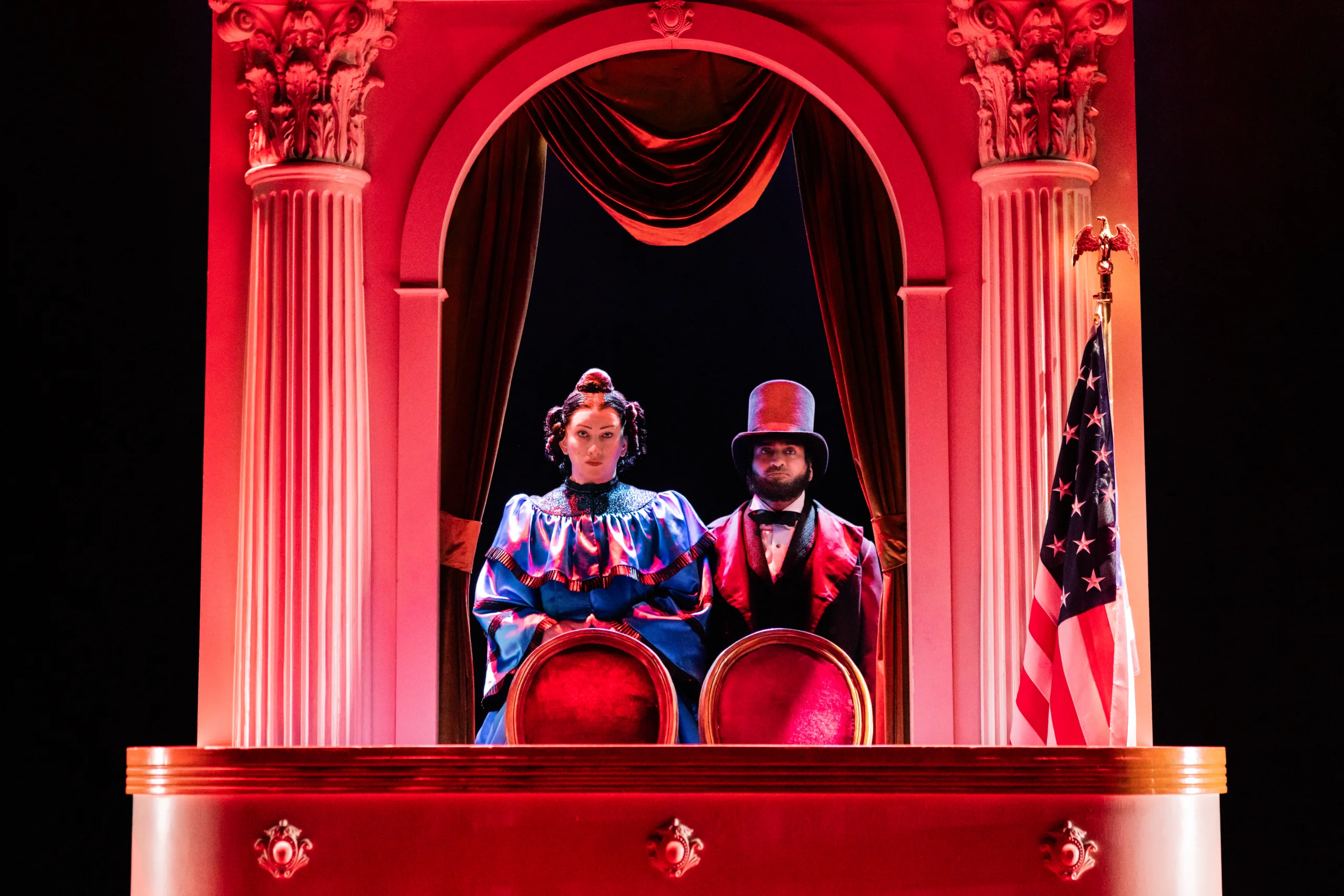 Two performers dressed in formal period costumes seated in an ornate theater box lit with dramatic red lighting, framed by tall columns and an American flag.