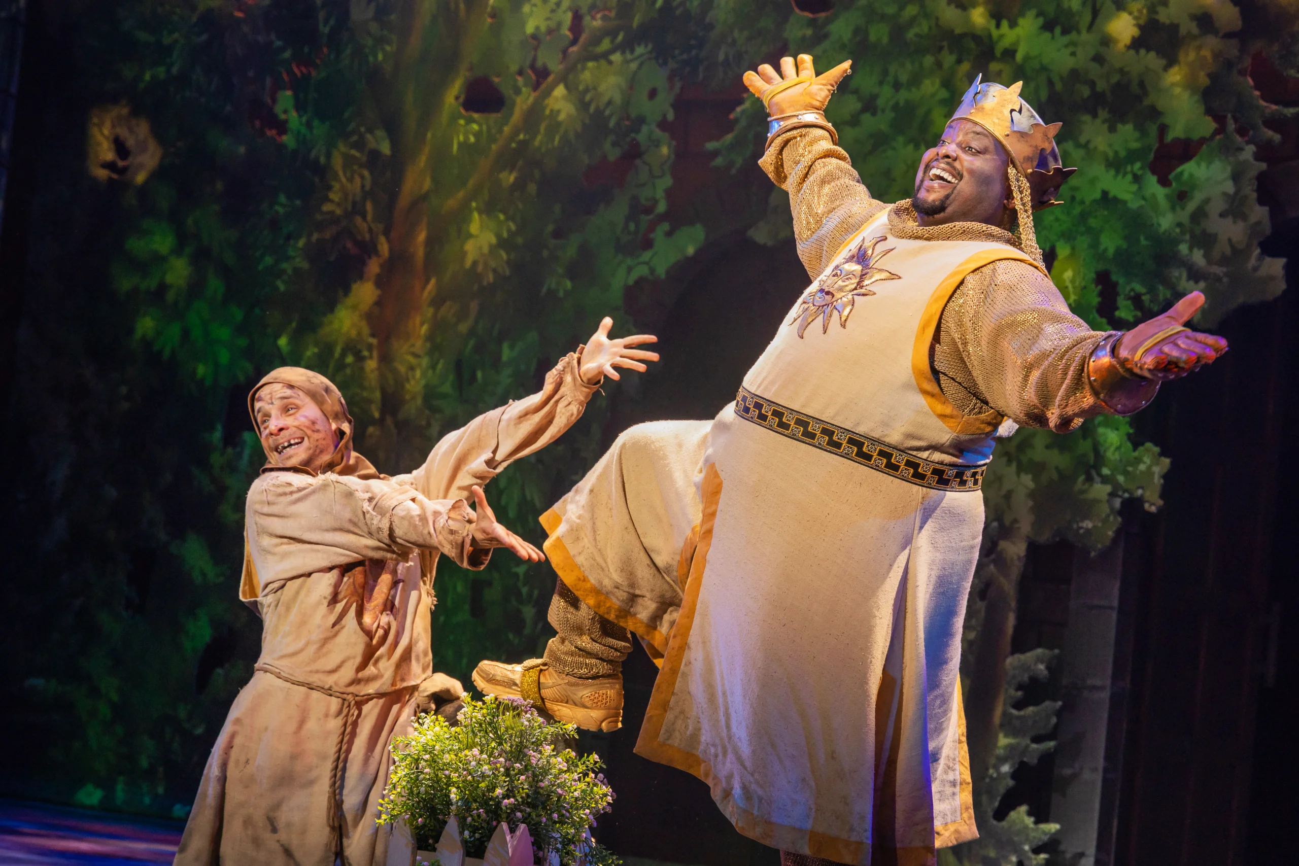 A costumed performer dressed as a king stands triumphantly onstage with arms outstretched while another robed performer reaches toward them during a lively theatrical scene set against a colorful forest backdrop.