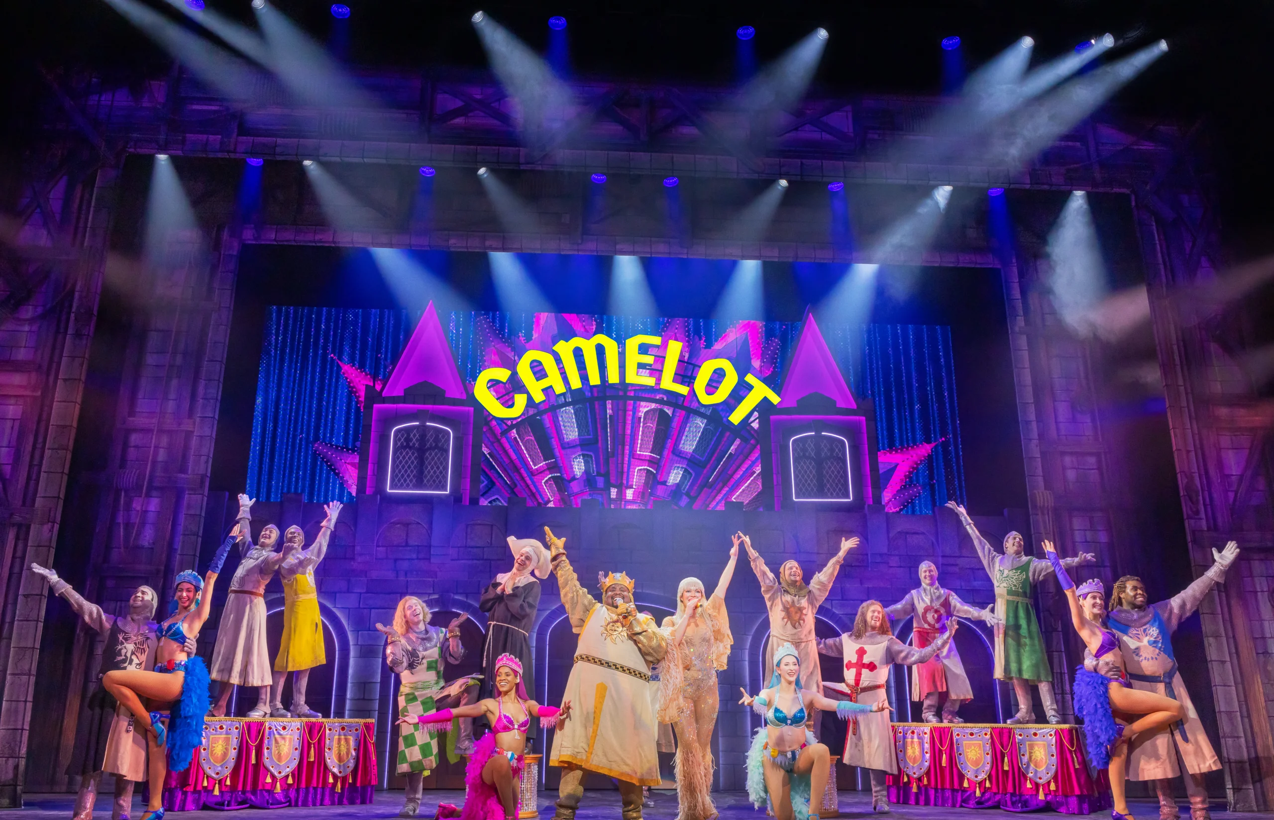 cast poses with outstretched hands above a Camelot sign.