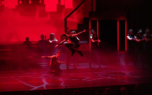 Contemporary dancers performing an energetic choreographed jump on a theater stage, illuminated by dramatic red lighting during a live stage production.