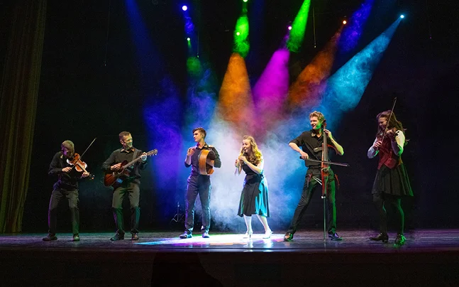 Live music ensemble performing on stage with violins, guitar, cello, and vocals under colorful theatrical lighting.