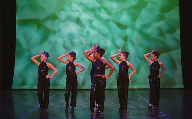 Contemporary dance performance featuring six dancers in black costumes on a professional theater stage with dramatic green lighting.