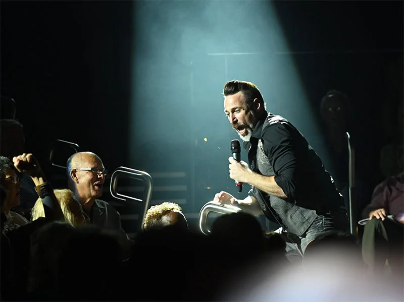 Rock singer performing live on stage, leaning toward audience with microphone during concert under spotlight.