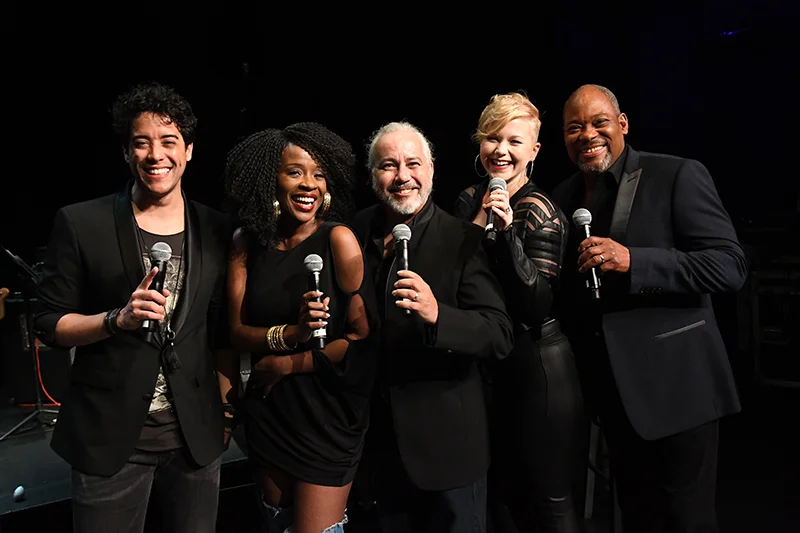 Five vocalists holding microphones posing together on stage during live rock and roll concert performance.