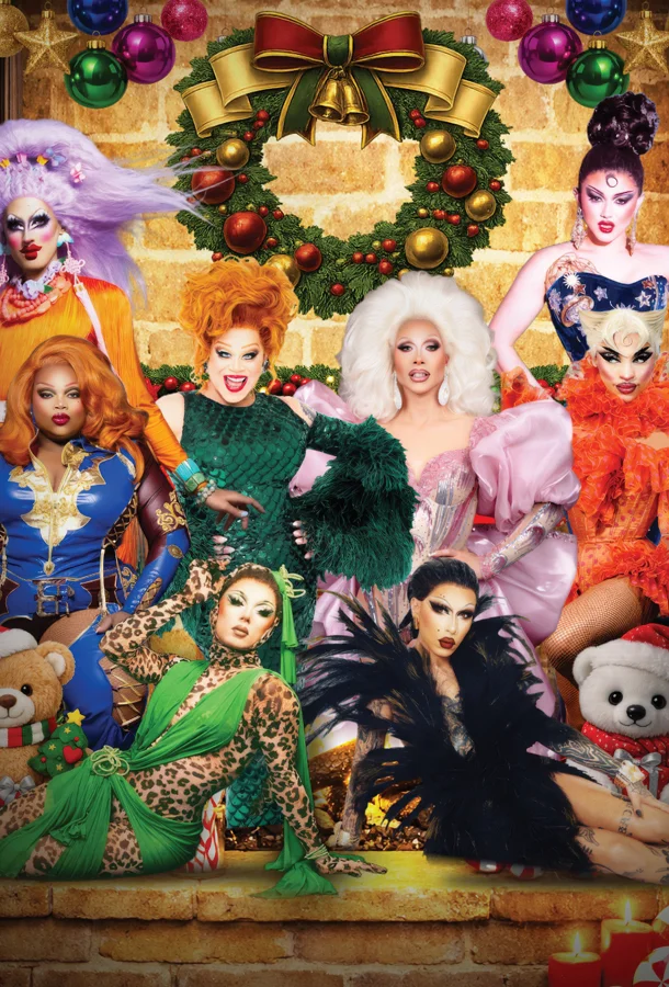 Group portrait of drag performers in colorful, glamorous costumes posing in front of a decorated Christmas wreath with ornaments, bows, and festive holiday decor on a brick wall background.