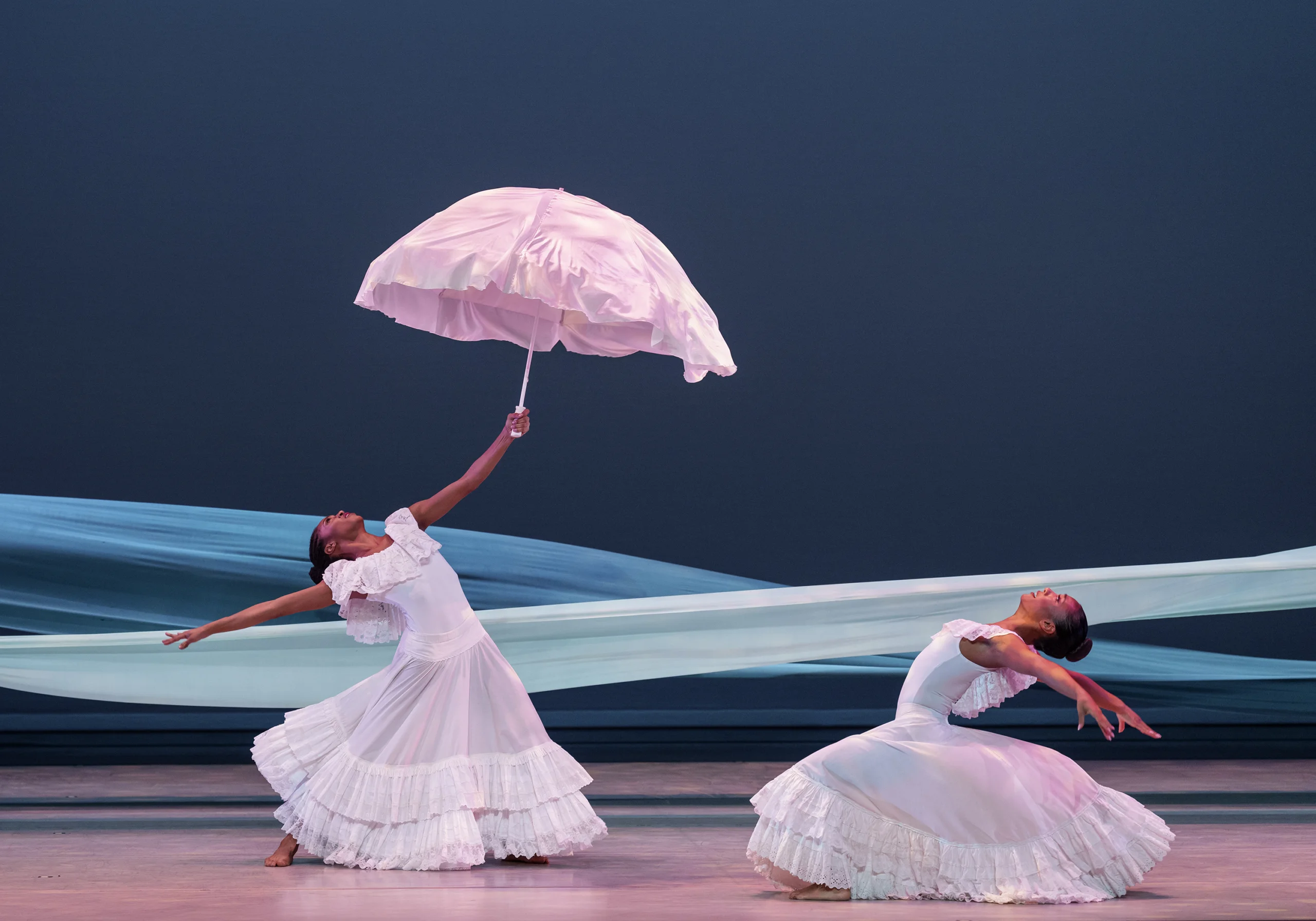 Credit Photo: ©Paul Kolnik
Two dancers in flowing white dresses perform with a parasol against a blue stage backdrop.