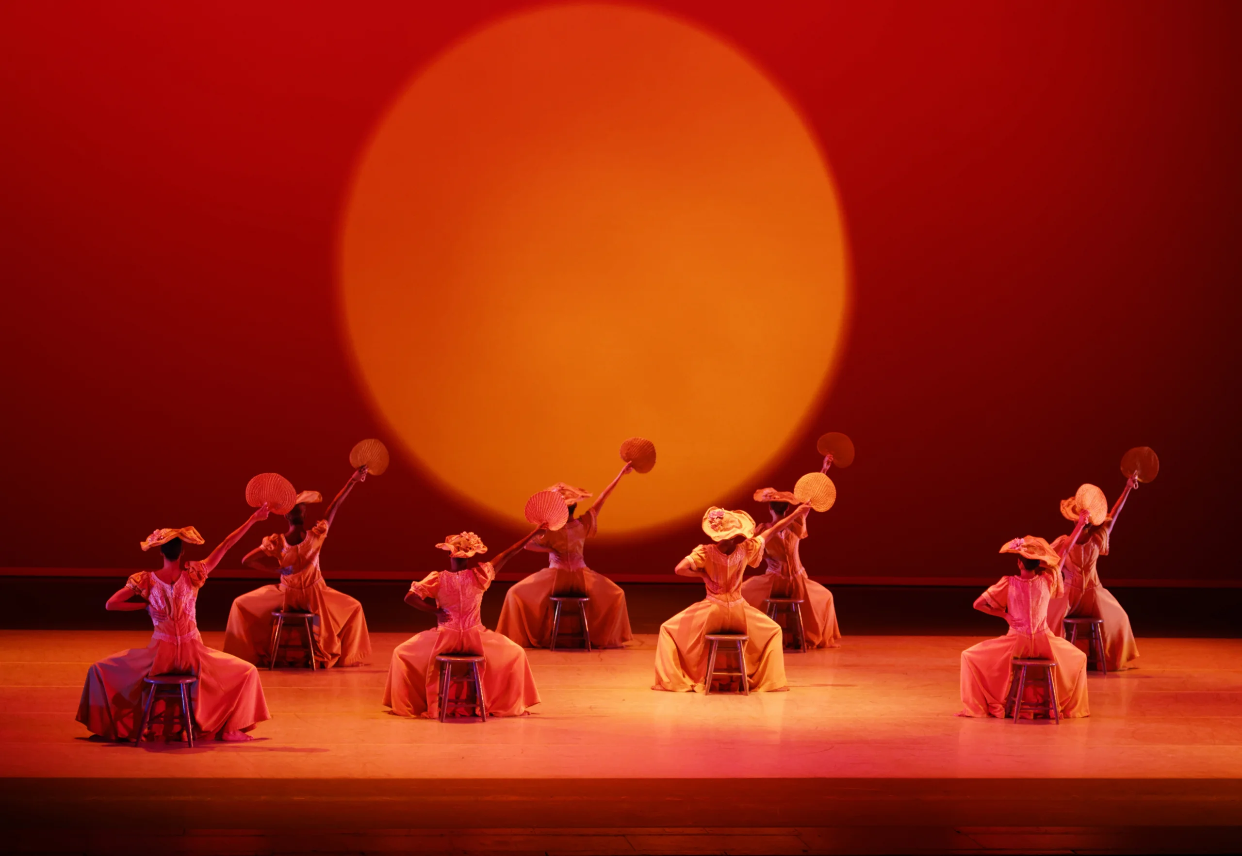 Line of dancers seated with fans raised on stage in front of a large glowing orange sun backdrop.