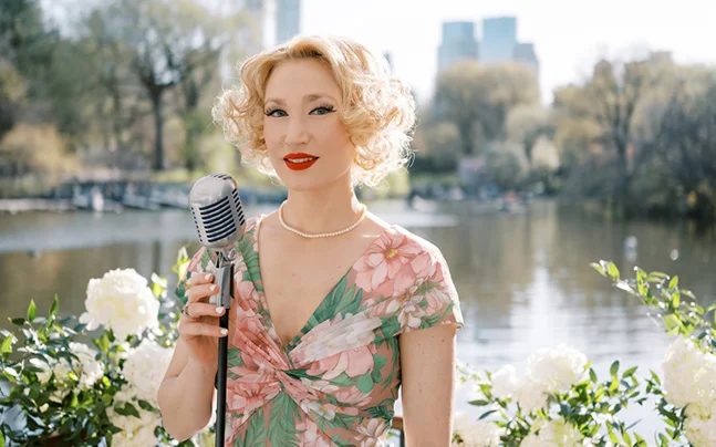 Performer in a floral dress holding a vintage microphone outdoors by a lake, representing a Doris Day tribute performance.